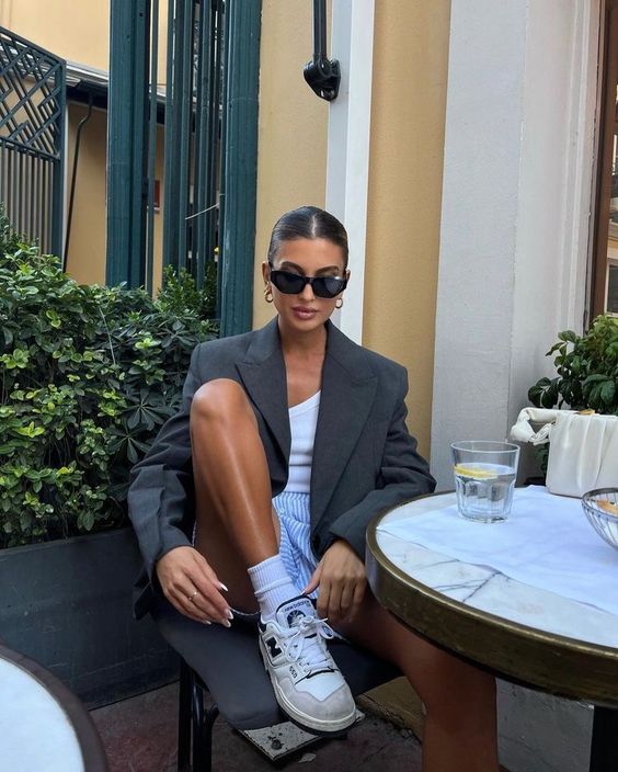 A woman exudes effortless style at a café, pairing a classic, oversized gray blazer with casual striped shorts. She completes the look with white crew socks, trendy white sneakers, and sleek black sunglasses. The ensemble is a chic blend of business and casual, perfect for a sunny day out.